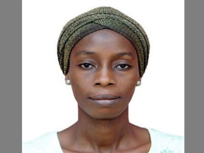 Eine Frau mit einem Kopftuch und Ohrringen blickt ruhig in die Kamera vor einem neutralen Hintergrund.