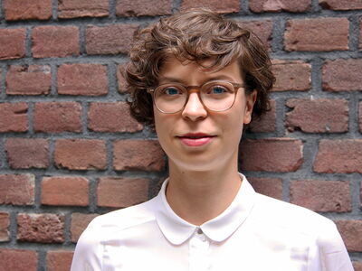 Eine Frau mit Brille und weißer Bluse steht lächelnd vor einer Ziegelwand.