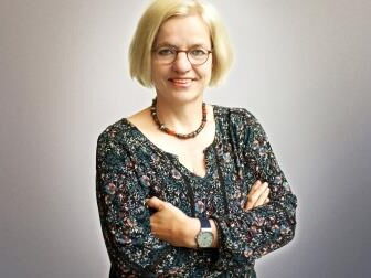 Eine Frau mit blonden Haaren und Brille lächelt selbstbewusst in die Kamera, die Arme vor der Brust verschränkt.