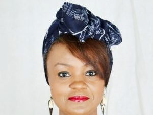 Eine Frau mit einem blauen Kopftuch und roten Lippen lächelt in die Kamera.