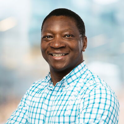 Ein Mann in kariertem Hemd lächelt selbstbewusst in die Kamera vor einem verschwommenen Hintergrund.