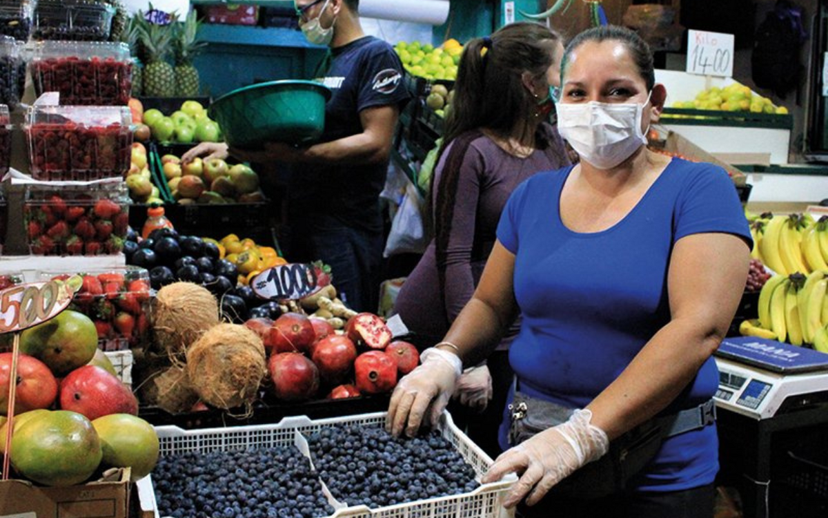 Eine Frau verkauft Blaubeeren auf einem lebhaften Markt, während sie einen Mundschutz und Handschuhe trägt.