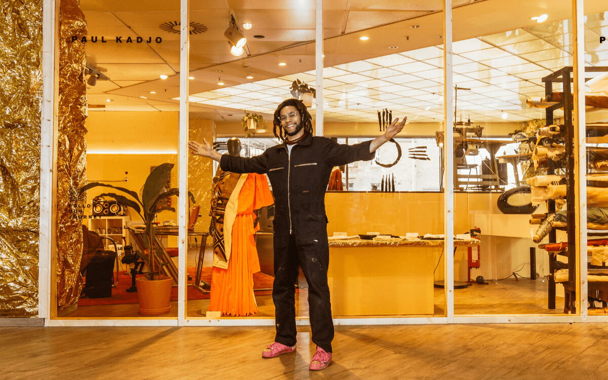 Ein Mann steht lächelnd mit ausgebreiteten Armen in einem kreativen Atelier voller Kunstwerke und Materialien.