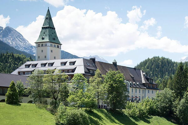 A grand building with a tall tower stands amidst lush greenery and mountains under a blue sky in Elmau.