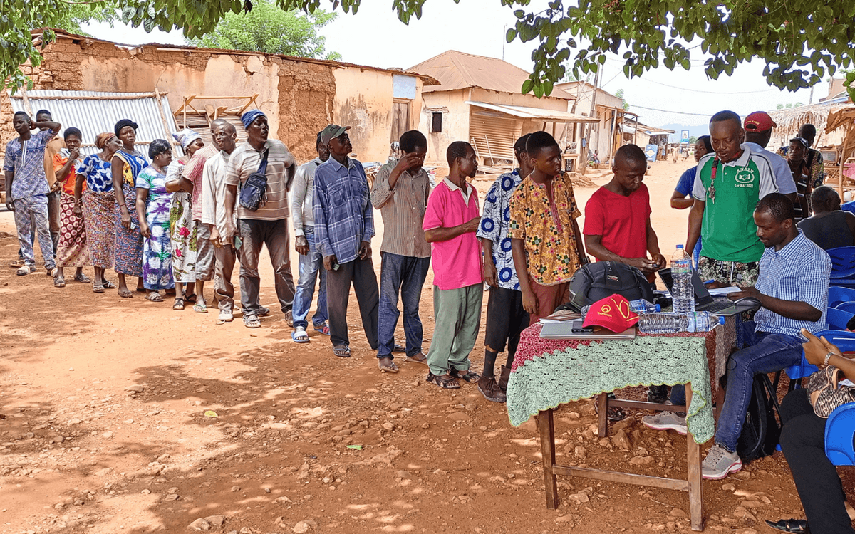 Menschen stehen in einer langen Schlange, um sich an einem Tisch im Freien registrieren zu lassen, während Gebana-Mitarbeiter assistieren.