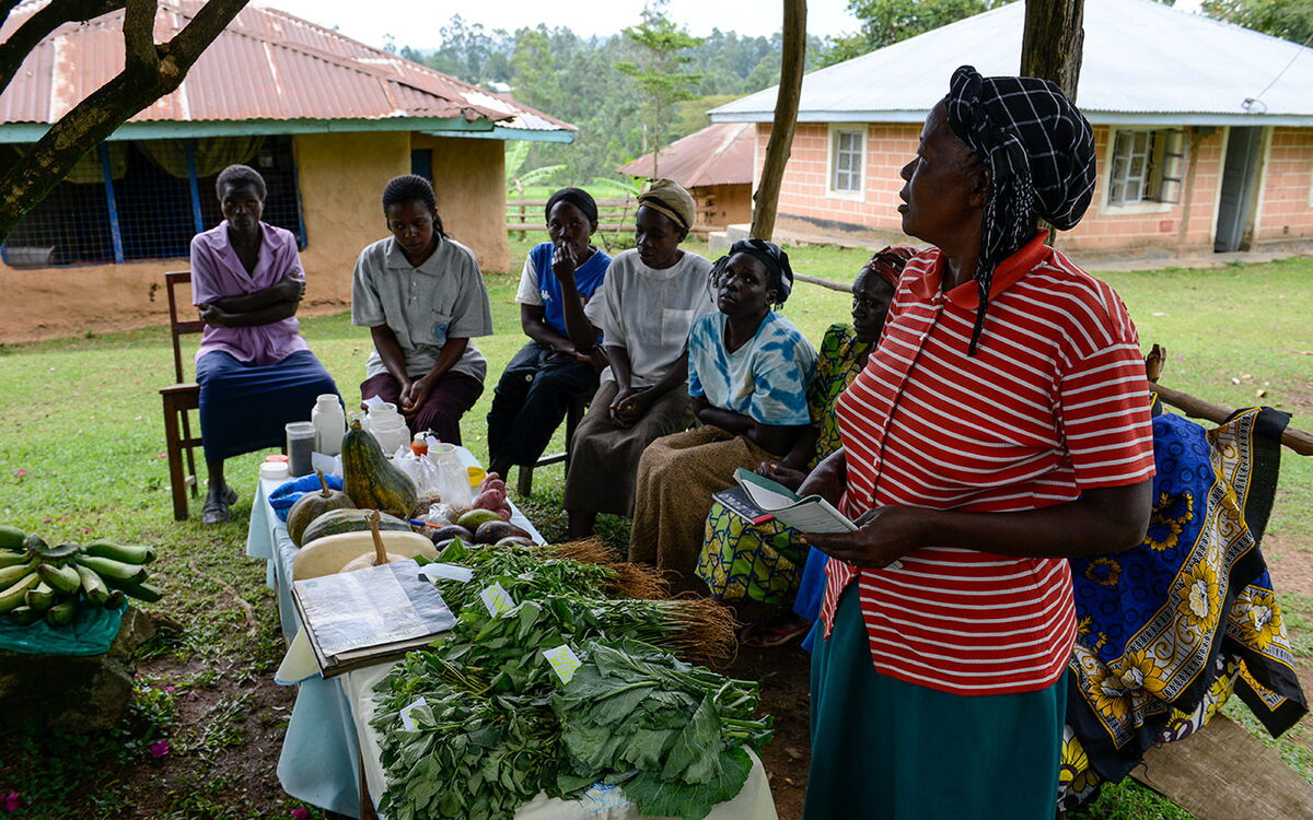 Eine Frau hält einen Vortrag über landwirtschaftliche Produkte vor einer Gruppe von Menschen auf einem Rasen vor einem Haus.