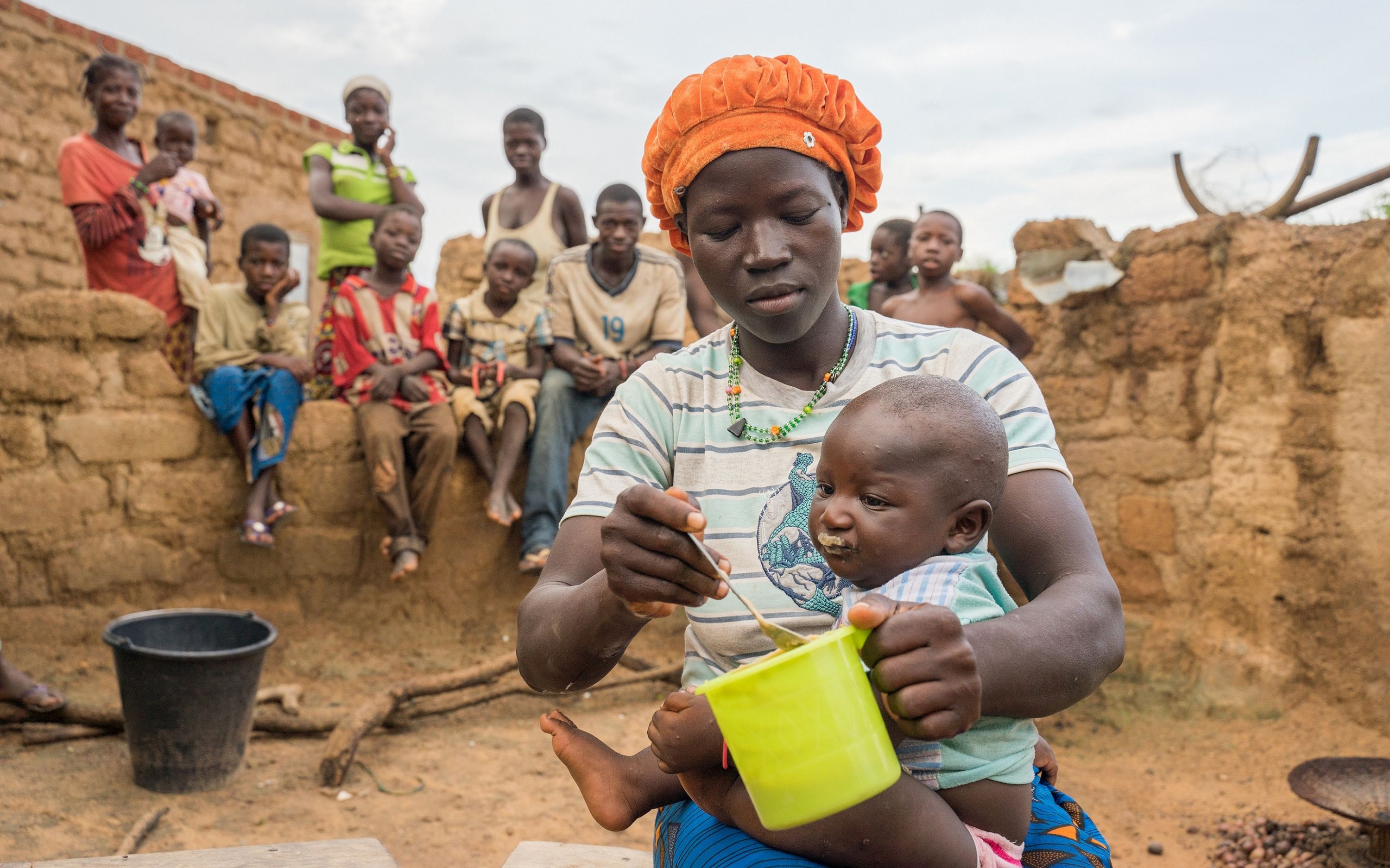Eine Frau in Burkina Faso füttert ein Kleinkind, während mehrere Kinder auf einer Mauer im Hintergrund sitzen.