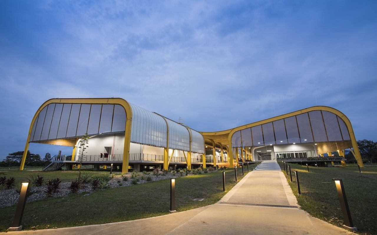 Ein modernes Gebäude mit geschwungenem Dach erstrahlt nachts in warmem Licht entlang eines beleuchteten Weges.