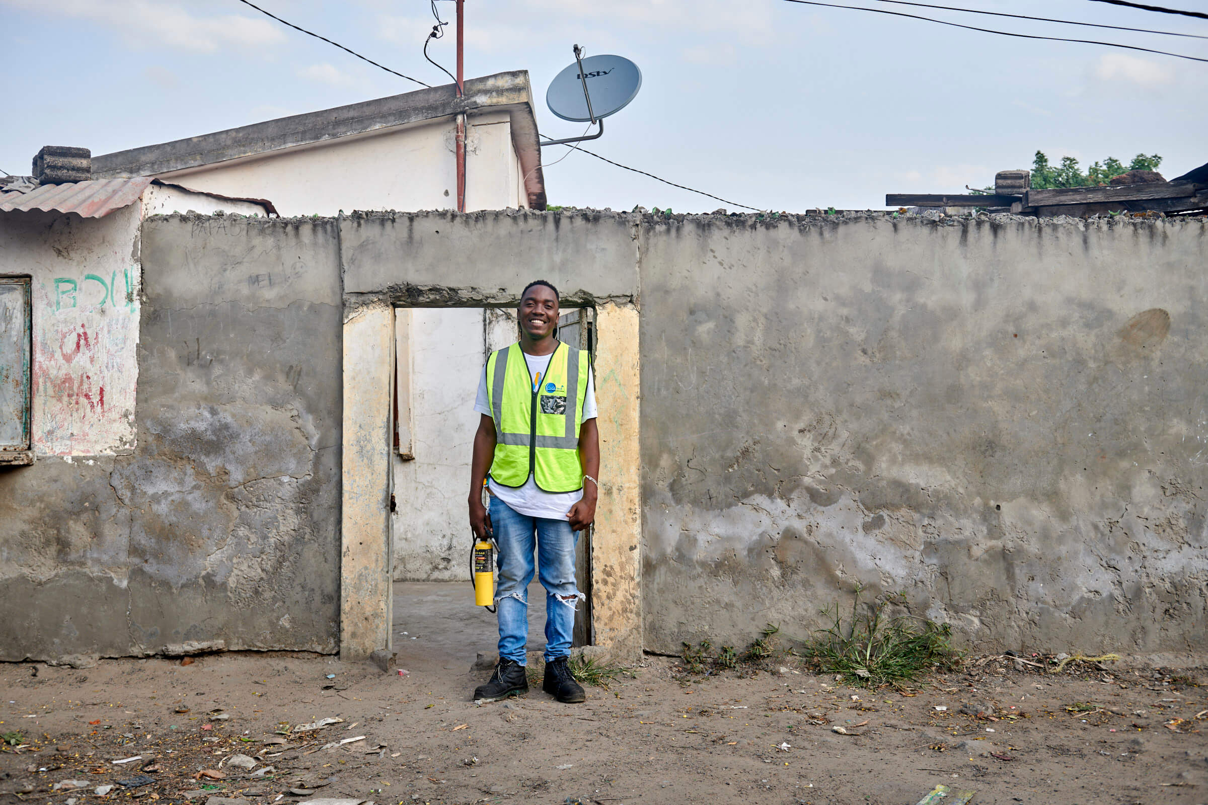 Ein Mann in einer gelben Weste steht lächelnd vor einem alten Haus mit einer Satellitenschüssel auf dem Dach.