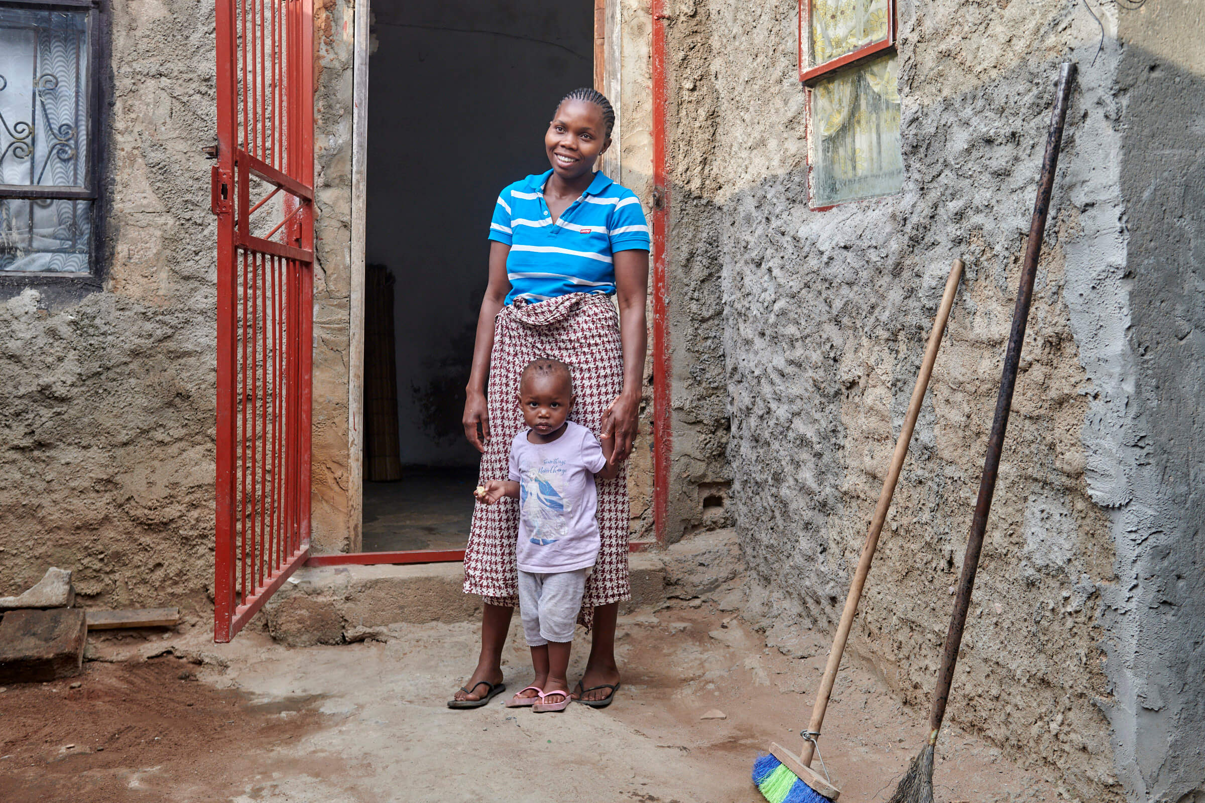 Minoca Adau Macucule steht lächelnd mit einem Kind vor ihrem Haus, umgeben von rustikalen Steinwänden.