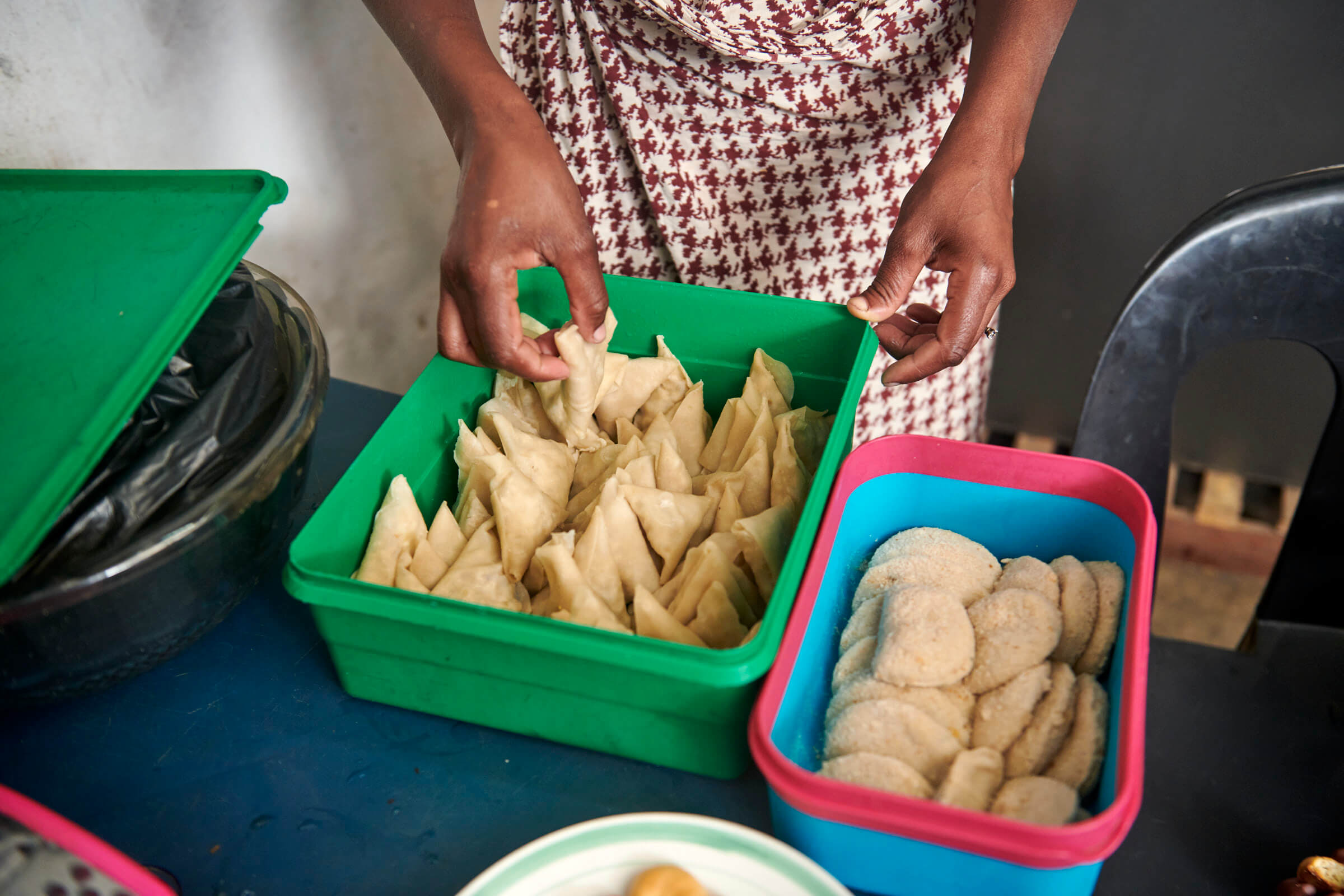 Eine Person legt Teigtaschen in eine grüne Box, während verschiedene Salgados in einer anderen Box aufbewahrt werden.