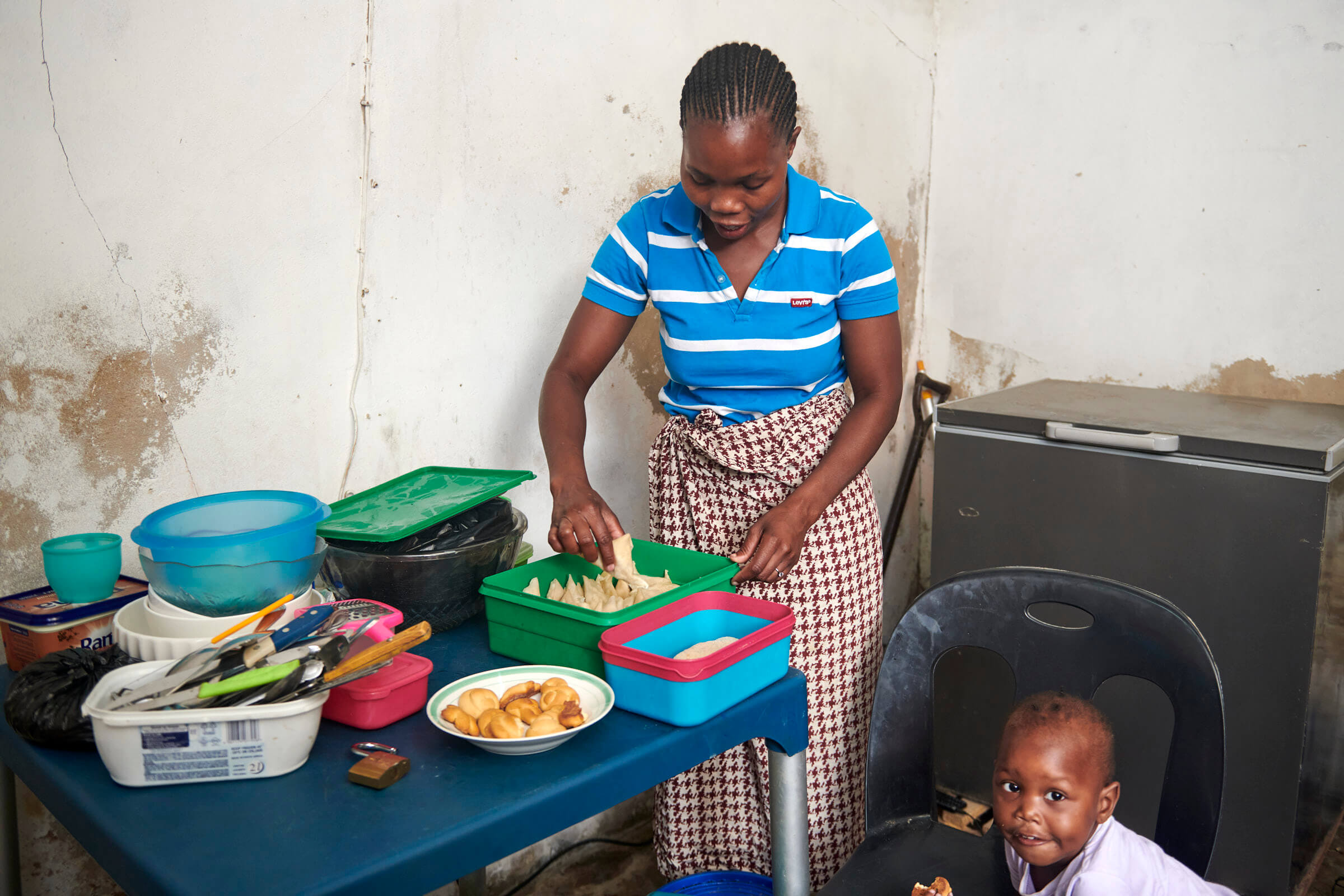 Eine Frau in gestreiftem Shirt bereitet in der Küche Essen vor, während ein Kind daneben auf einem Stuhl sitzt.