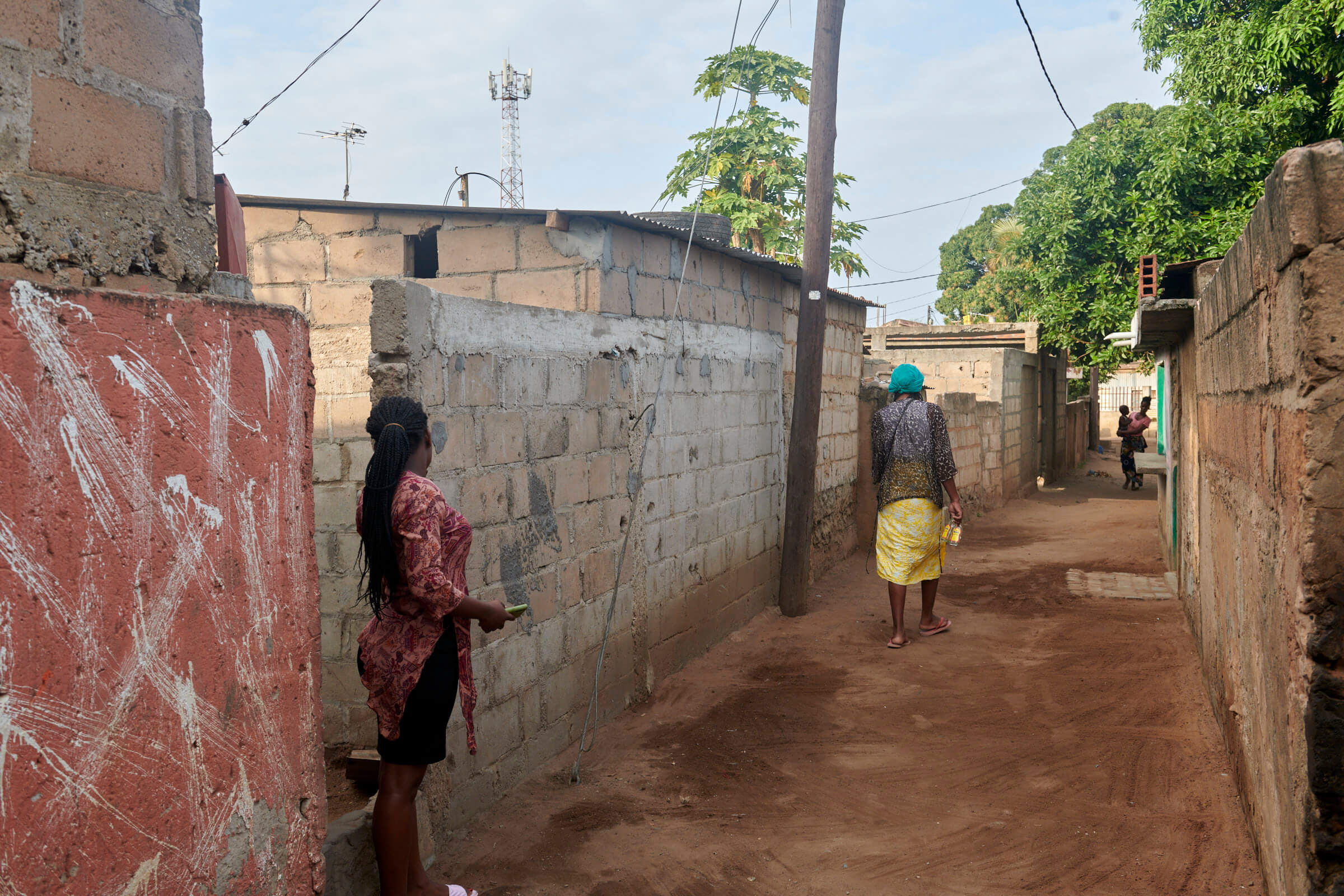Eine Frau in bunter Kleidung geht durch eine schmale Gasse in Macucules Nachbarschaft, umgeben von Steinmauern und Bäumen.