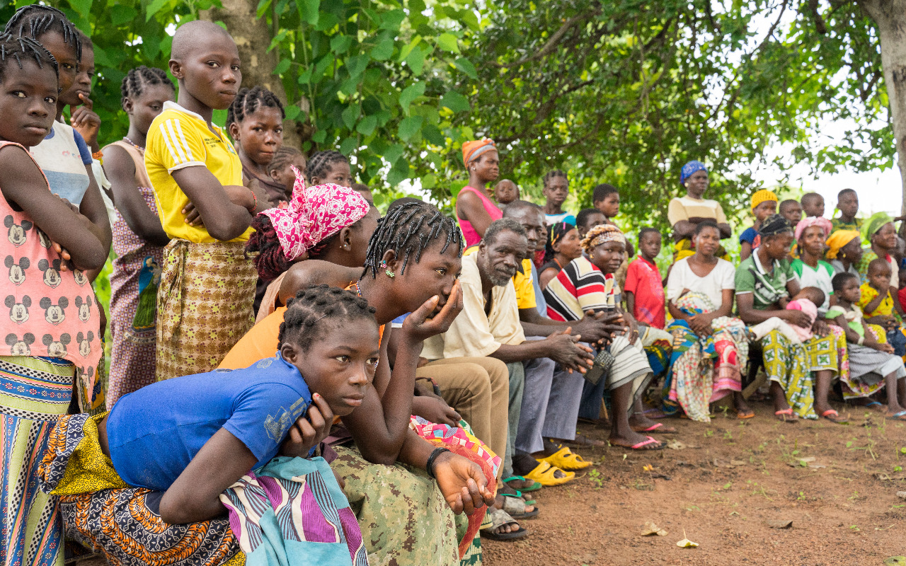 Eine Gruppe von Menschen versammelt sich im Freien unter einem Baum in Burkina Faso, um einer Präsentation zuzuhören.