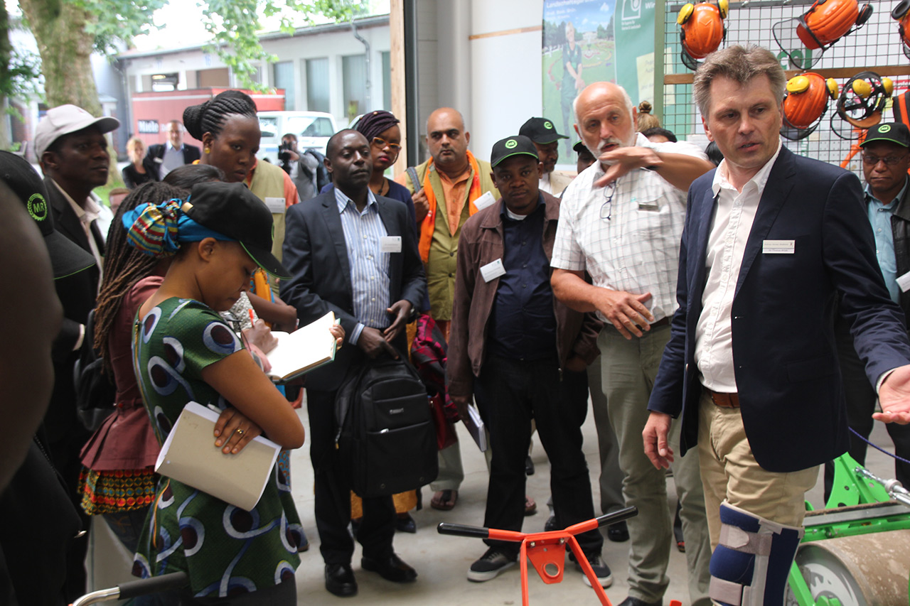 Eine Gruppe von Menschen lauscht einem Vortrag über landwirtschaftliche Geräte in einem Ausstellungsraum.