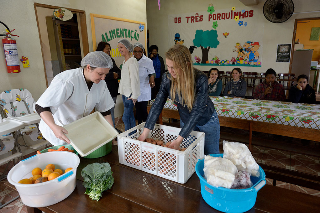 Zwei Frauen sortieren Lebensmittel in einer Kita, während Kinder im Hintergrund an einem Tisch sitzen.