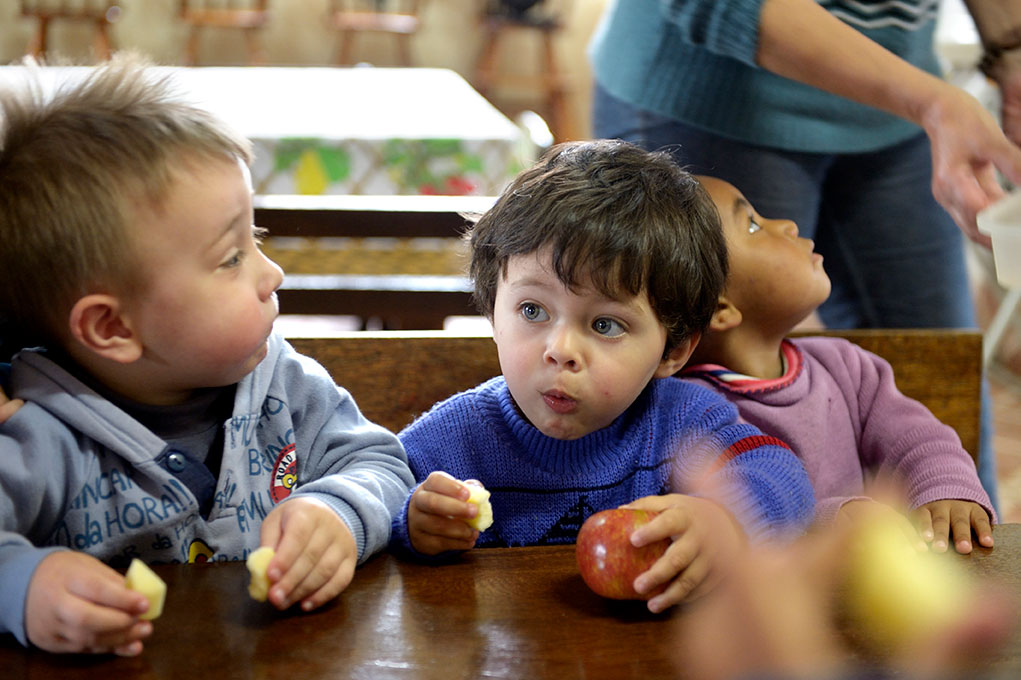 Drei Kinder sitzen an einem Tisch und essen Äpfel, während sie neugierig in verschiedene Richtungen schauen.