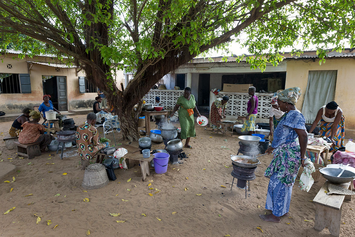 Frauen kochen gemeinsam unter einem großen Baum in einem sandigen Hof vor Gebäuden, umgeben von Töpfen und Kochutensilien.