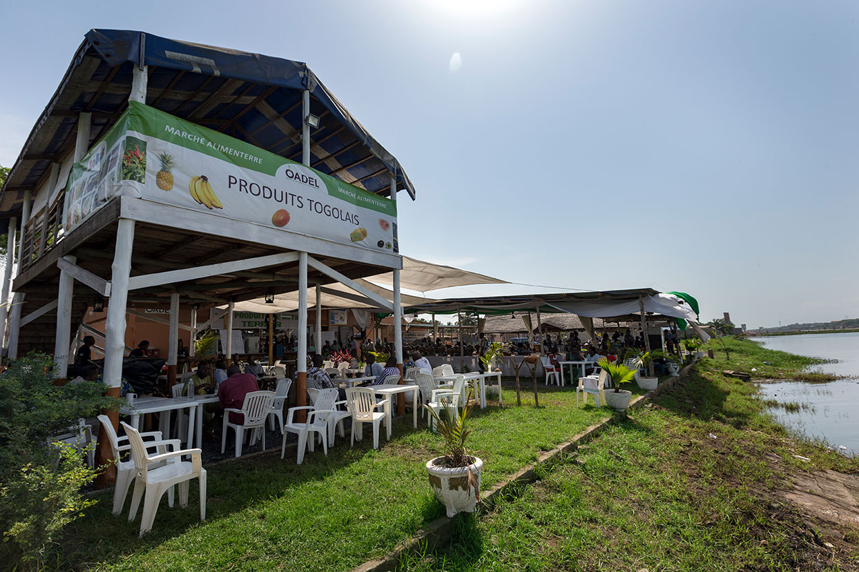 Menschen sitzen unter einem überdachten Bereich eines togoischen Marktes am Flussufer und genießen die lokale Atmosphäre.