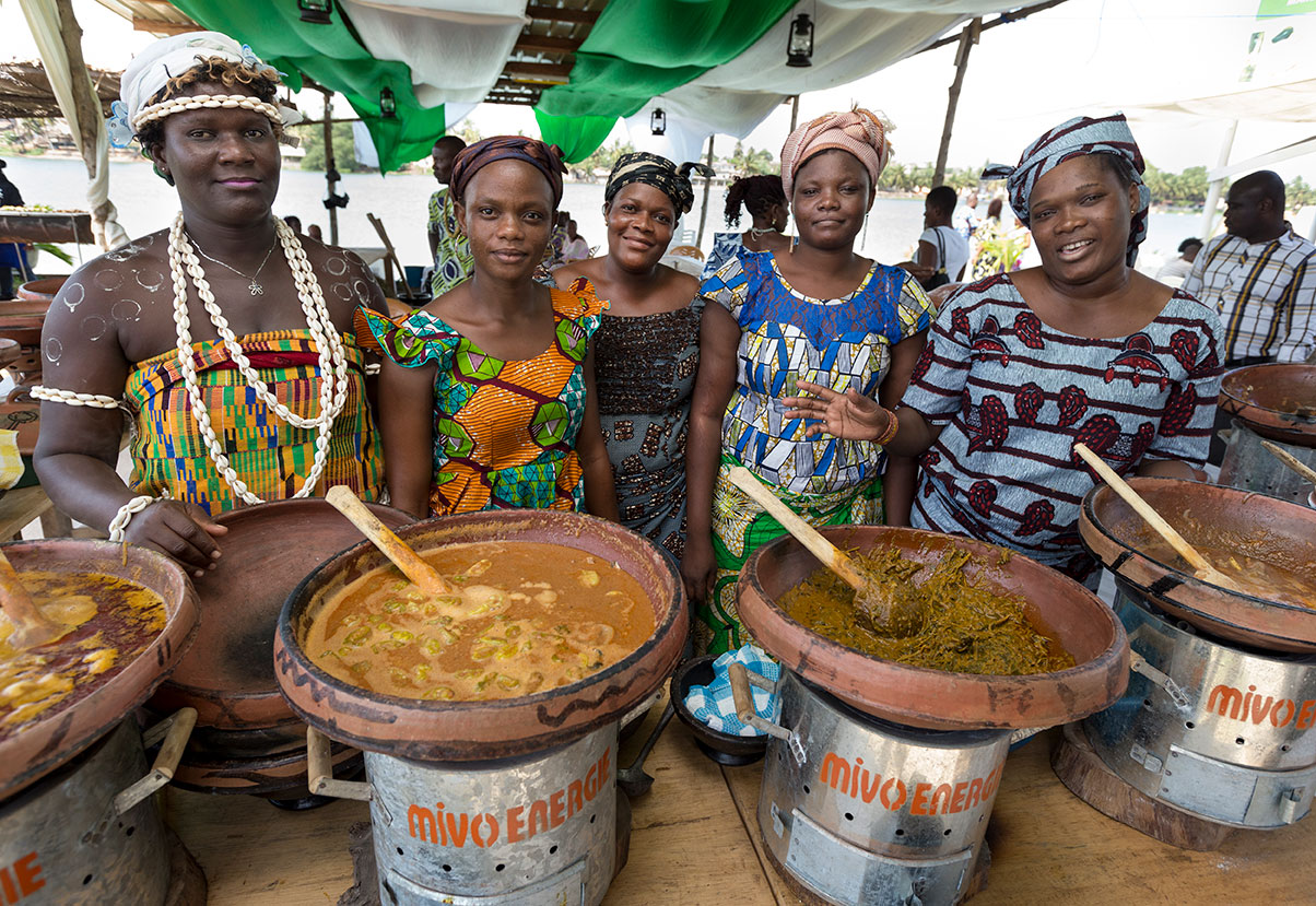 Fünf Frauen in bunter Kleidung stehen lächelnd hinter traditionell gekochtem Essen auf einem Markt.