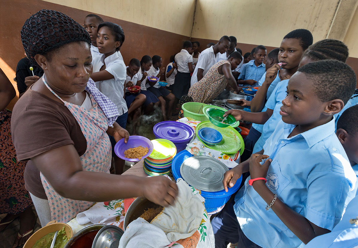 Eine Frau serviert Essen an Schüler in Schuluniformen, die in einer Kantine geduldig mit Tellern warten.