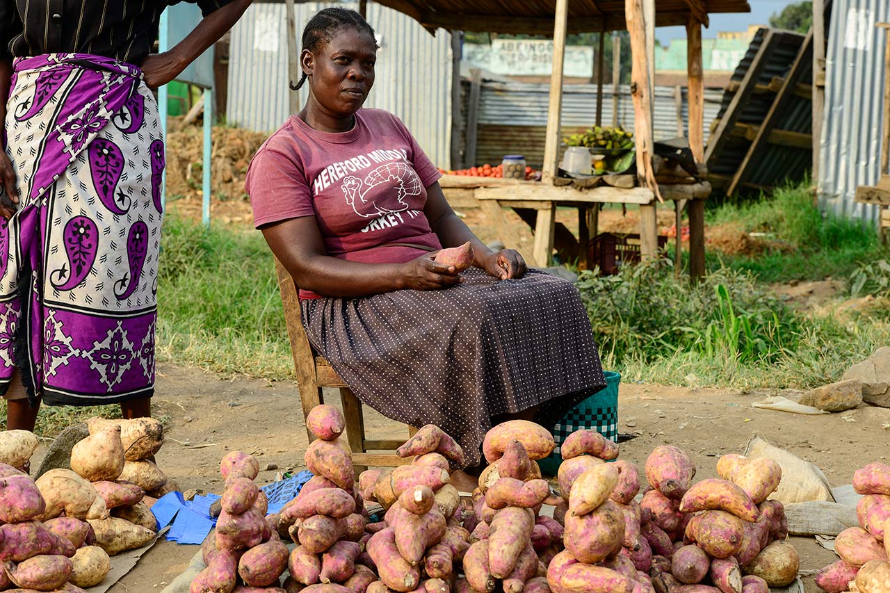 A woman in Kenya sits by a market stall displaying piles of sweet potatoes for sale.
