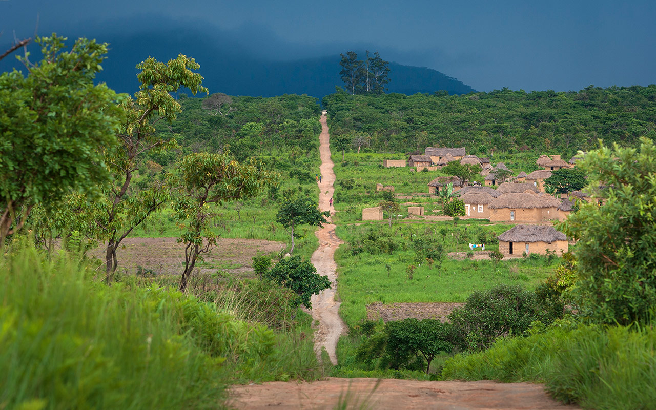 A narrow dirt path leads to a village with thatched-roof huts surrounded by lush greenery and a dark, cloudy sky.