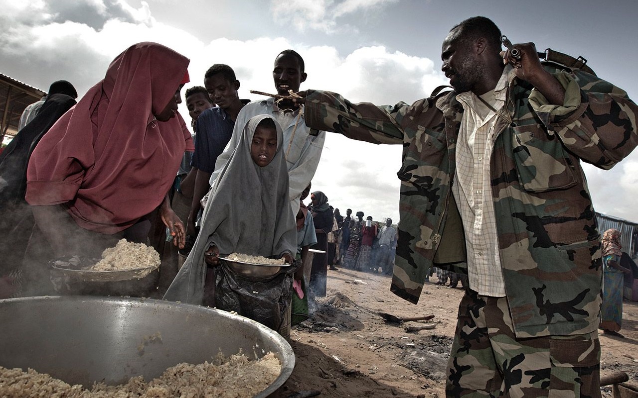 Ein Mann in Tarnkleidung überwacht die Essensverteilung an eine Gruppe von Menschen, darunter Kinder und Erwachsene.